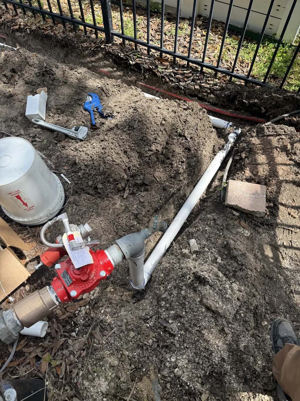 New valve being plumbed into an irrigation line in an open trench beside a fence